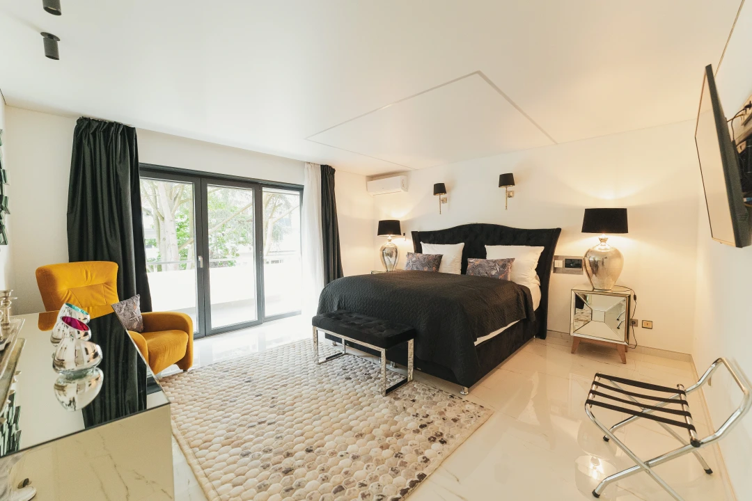 Dieses Bild zeigt das Schlafzimmer mit dem schwarzen Bett von der Eingangstüre aus. Es befindet sich ein Teppich, ein orangener Sessel, eine Komode sowie ein an der Wand hängender Flachbild-TV an der Wand. Das Zimmer hat einen Zugang zu einem Balkon.  
