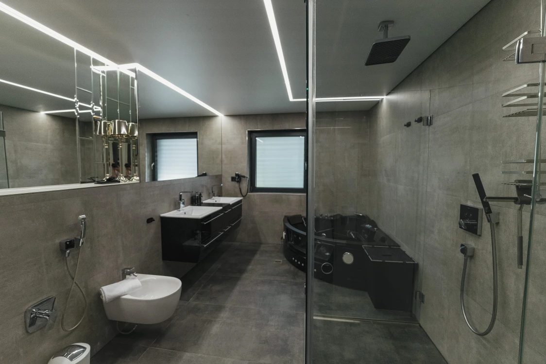 Großes Bad mit grauen Fliesen, Dusche, 2 Waschbecken, großer Spiegel, Toilette, Badewanne mit Jacuzzi.