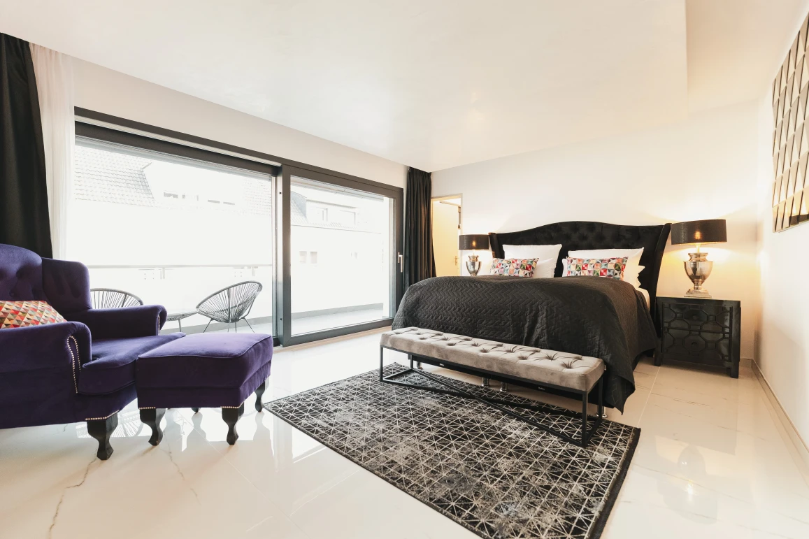 Dieses Bild zeigt das Bett mit den bunt karrierten Kissen. Nun befindet sich ein lilaner Sessel im Bild, welcher sich in der Ecke des Raumes befindet.