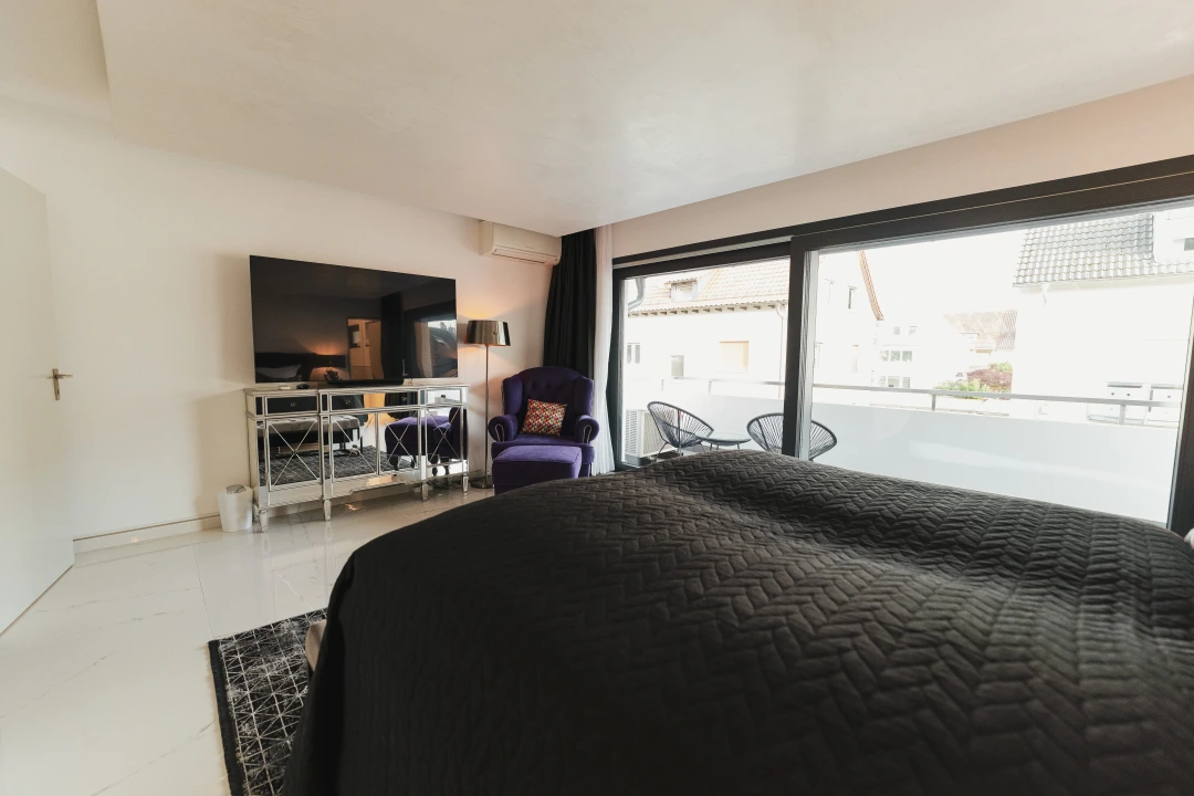 Auf diesem Bild befindet sich ein weiteres Schlafzimmer mit einem Doppelbett auf welchem eine schwarze Bettdecke ist. Ein lilaner Sessel sowie ein Flachbild-TV auf einer Komode, sind im Hintergund zu erkennen. Auch dieses Zimmer verfügt über einen Balkon.