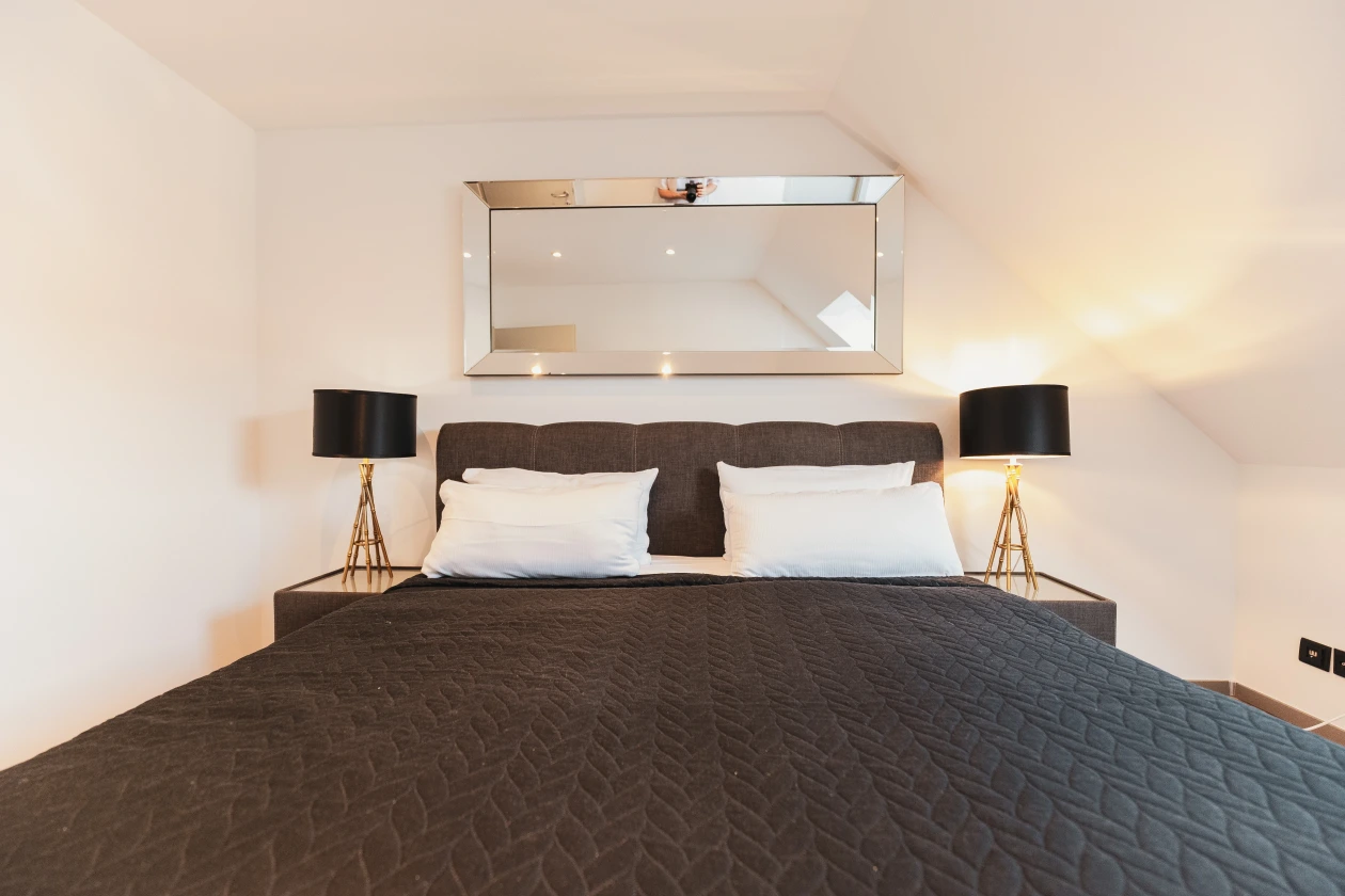 Das Bild zeigt ein graues Doppelbett Bett mit einer schwarzen Decke, zwei Nachttischlampen und einem Spiegel über dem Bett. Das Zimmer hat eine Dachschräge.