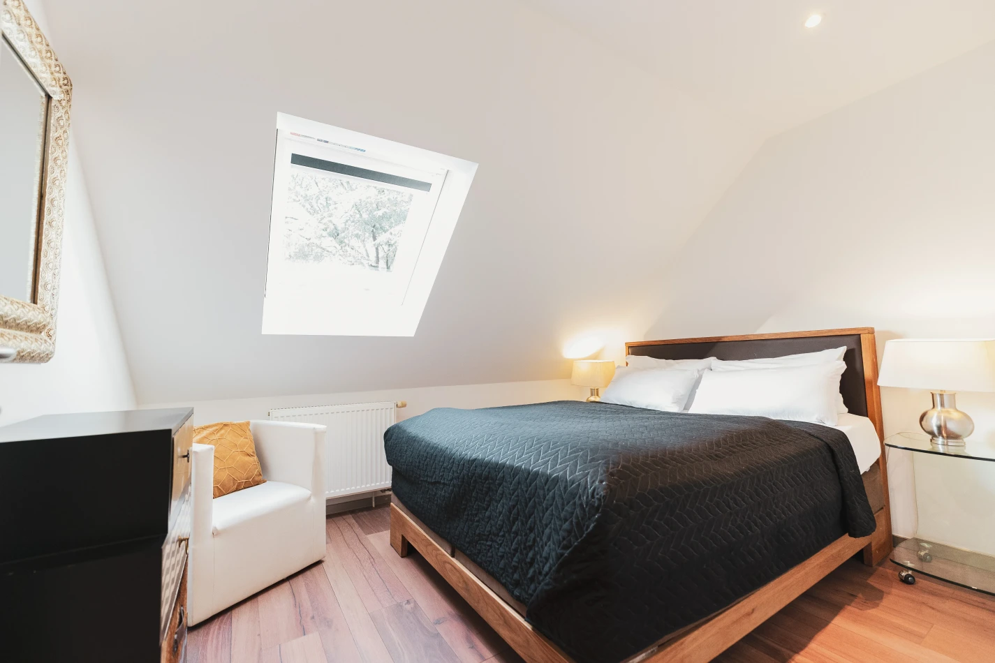 Dieses Bild zeigt ein Doppelbett aus Holz mit einer schwarzen Bettdecke. Der Raum hat eine Dachschräge, eine schwarze Komode, einen Spiegel sowie einen weißen Sessel.