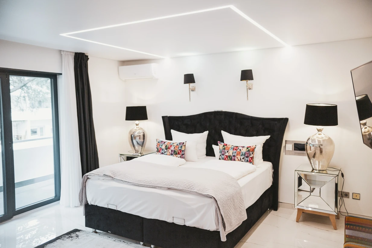 Dieses Bild zeigt ein weiteres Doppelbett mit bunt karierten Kissen und weißem Bettbezug. Auch hier befindet sich zwei Nachttischlampen und ein Flachbild-TV an der Wand.